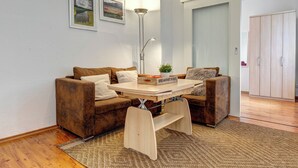 Un salon chaleureux avec un canapé marron, une table basse et une lampe. Deux tableaux encadrés ornent le mur, et une porte est visible au fond.