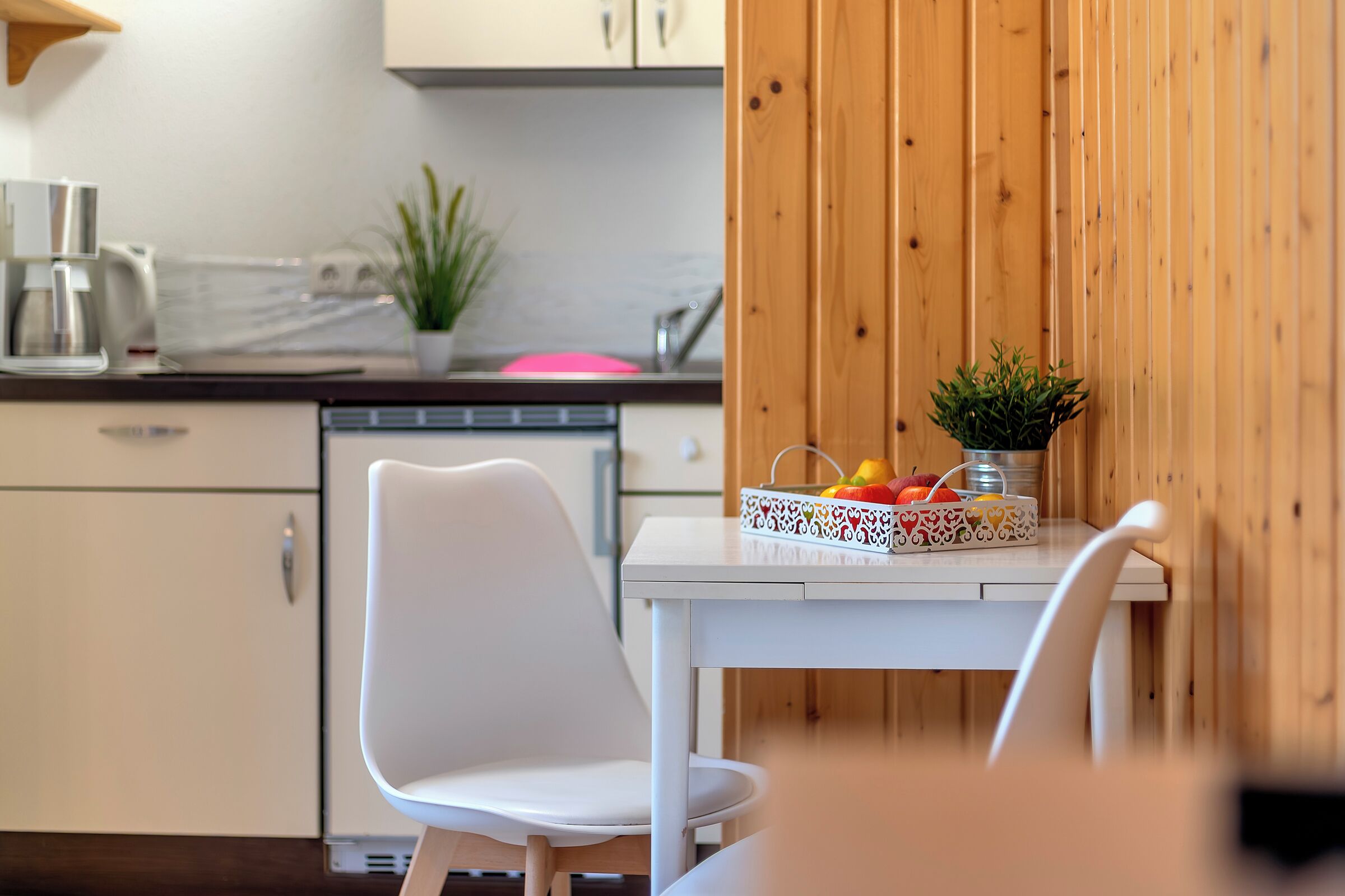 A modern kitchen with white cabinets, a wooden wall, a white table with a fruit basket, and a white chair.