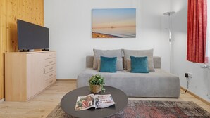 Una sala de estar moderna con un sofá, una mesa de centro con revistas, un televisor sobre un mueble de madera y un cuadro con una escena de playa en la pared.