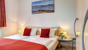 Et hotelværelse med en seng, et sengebord med en lampe, et vægmonteret tv og et billede af en strand.