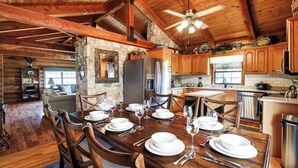 A wooden cabin interior with a dining area set for a meal, a stone fireplace, and a kitchen with wooden cabinets and a stone wall. - Canyon Lake Haven Cabin Hot Tub Yard BBQ Fire Pit (Canyon Lake)