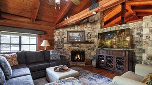 A cozy log cabin interior with a stone fireplace, wooden beams, a sofa, and a large window. - Canyon Lake Haven Cabin Hot Tub Yard BBQ Fire Pit (Canyon Lake)