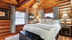 A log cabin bedroom with a large bed, two bedside tables with lamps, a bench, and a window with blinds. - Canyon Lake Haven Cabin Hot Tub Yard BBQ Fire Pit (Canyon Lake)