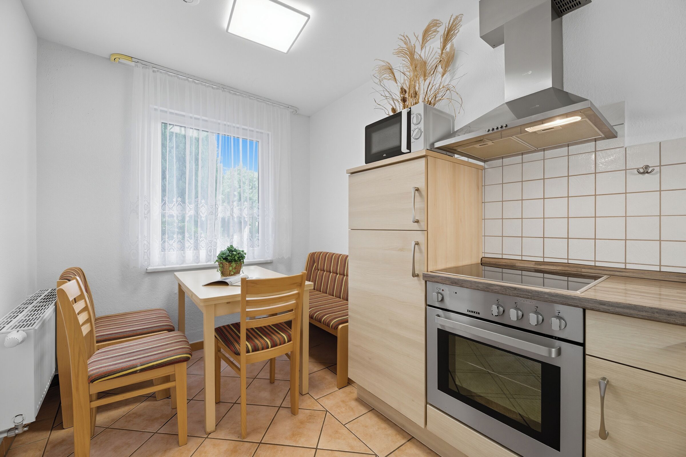Eine kompakte Küche mit Einbauofen, Mikrowelle und Herd. Es gibt einen Esstisch mit Stühlen und ein Fenster mit Vorhängen.