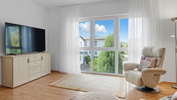 Un soggiorno moderno con una TV a schermo piatto su un mobile in legno, una poltrona beige con un cuscino e un'ampia finestra che si affaccia su edifici e spazi verdi.