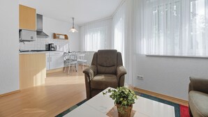 A modern living room with a brown leather armchair, a glass coffee table with a plant, and a view of a kitchen and dining area through sheer curtains.