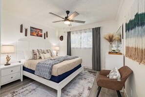 Ein Schlafzimmer mit Bett, Nachttischen, Deckenventilator, Stuhl und einem Fenster mit Vorhängen.