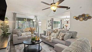 Ein Wohnzimmer mit einer Couch, Couchtisch und Dekokissen. Ein Deckenventilator und Wanddekorationen sind vorhanden.