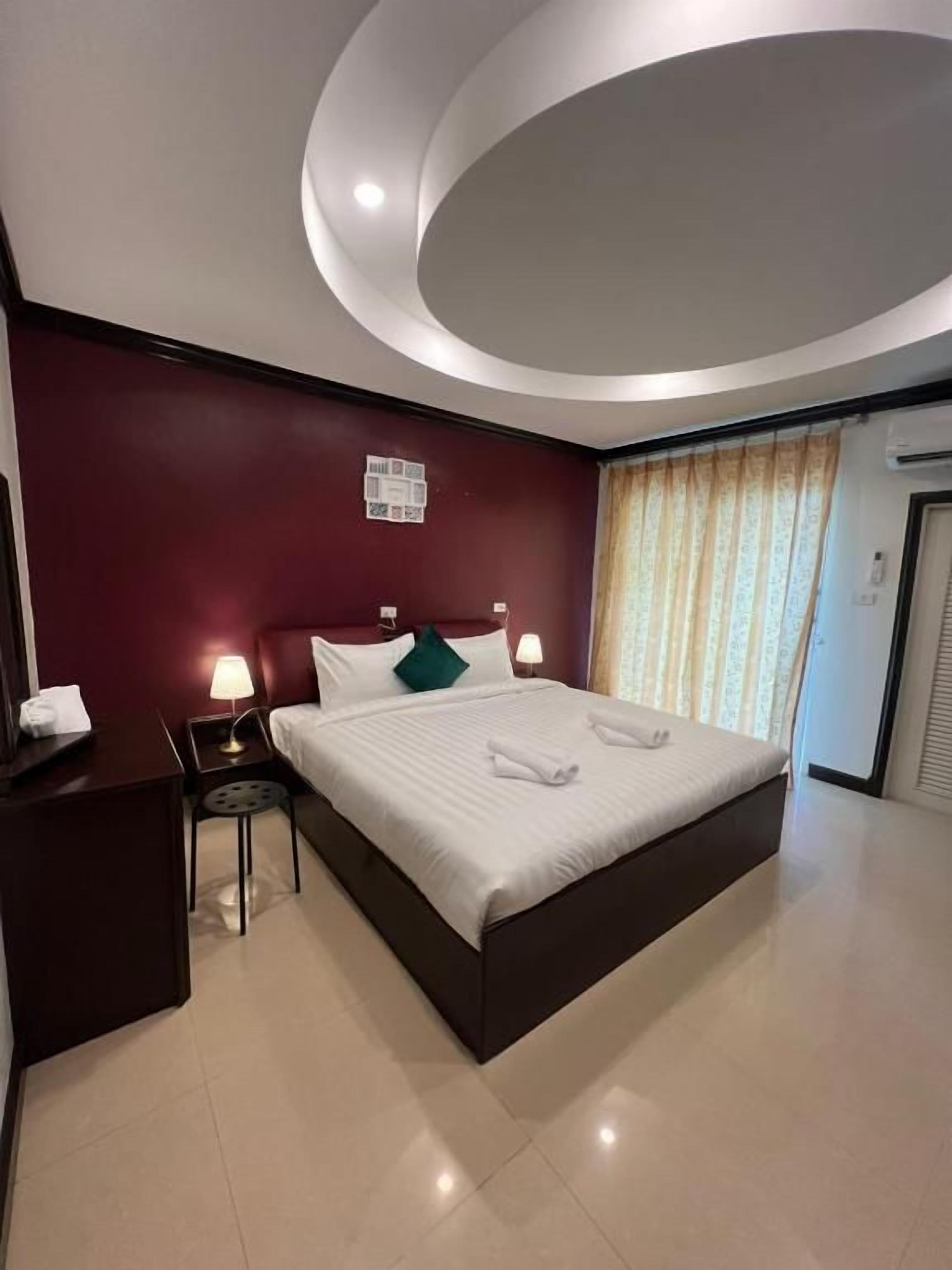 A modern hotel room with a large bed, two bedside tables with lamps, a dark-colored headboard, a red accent wall, and a large window with curtains.