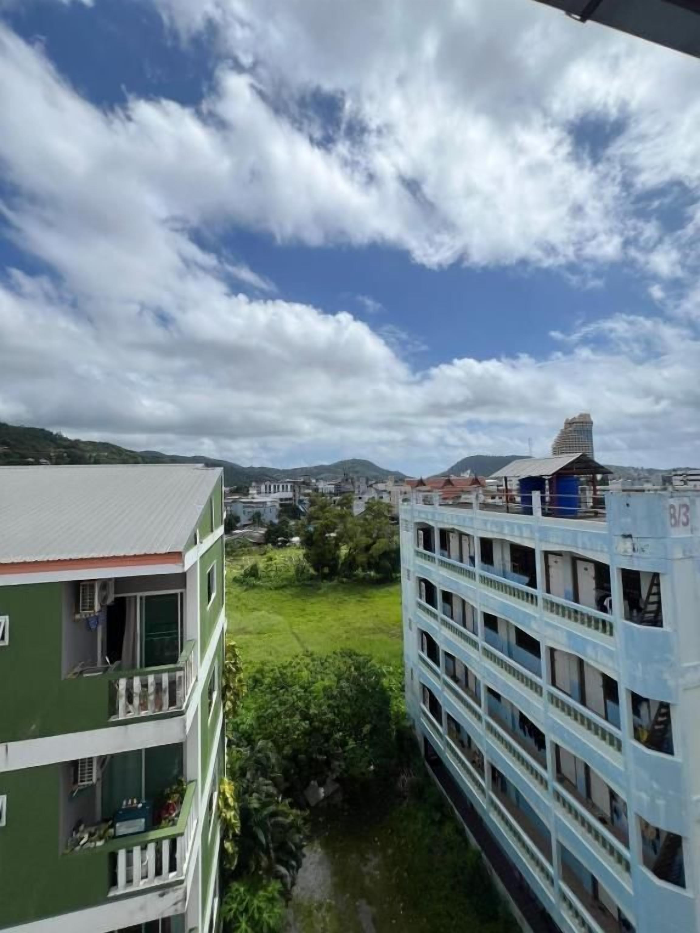 Un edificio de varios pisos con balcones, un edificio verde contiguo y una vista de un paisaje urbano lejano bajo un cielo parcialmente nublado.