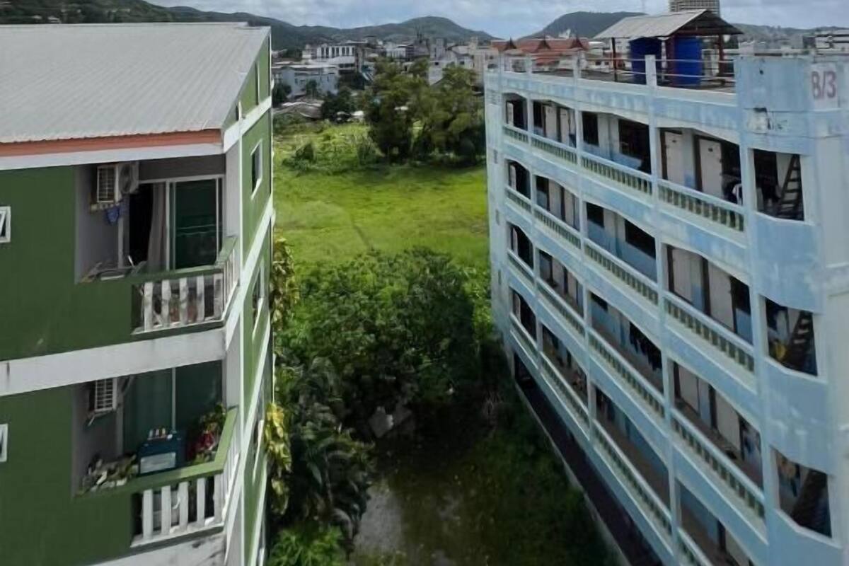 Um edifício com várias andares e varandas, um edifício verde adjacente a ele, e vista de um horizonte urbano distante sob um céu parcialmente nublado.