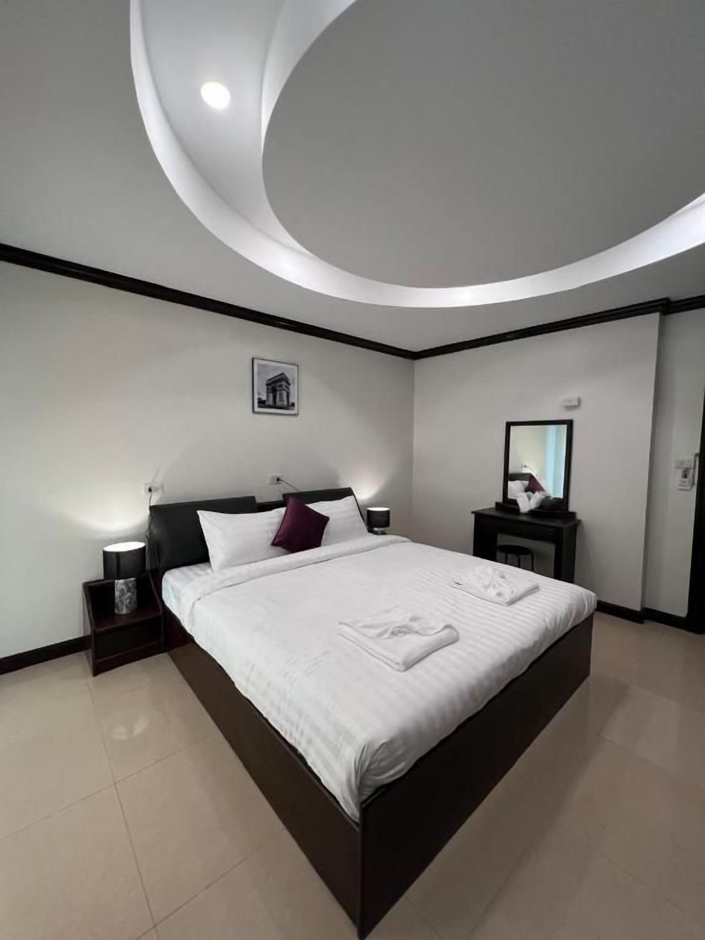 A modern hotel room with a large bed, a nightstand, a bedside table, a wall-mounted mirror, and a ceiling light fixture.
