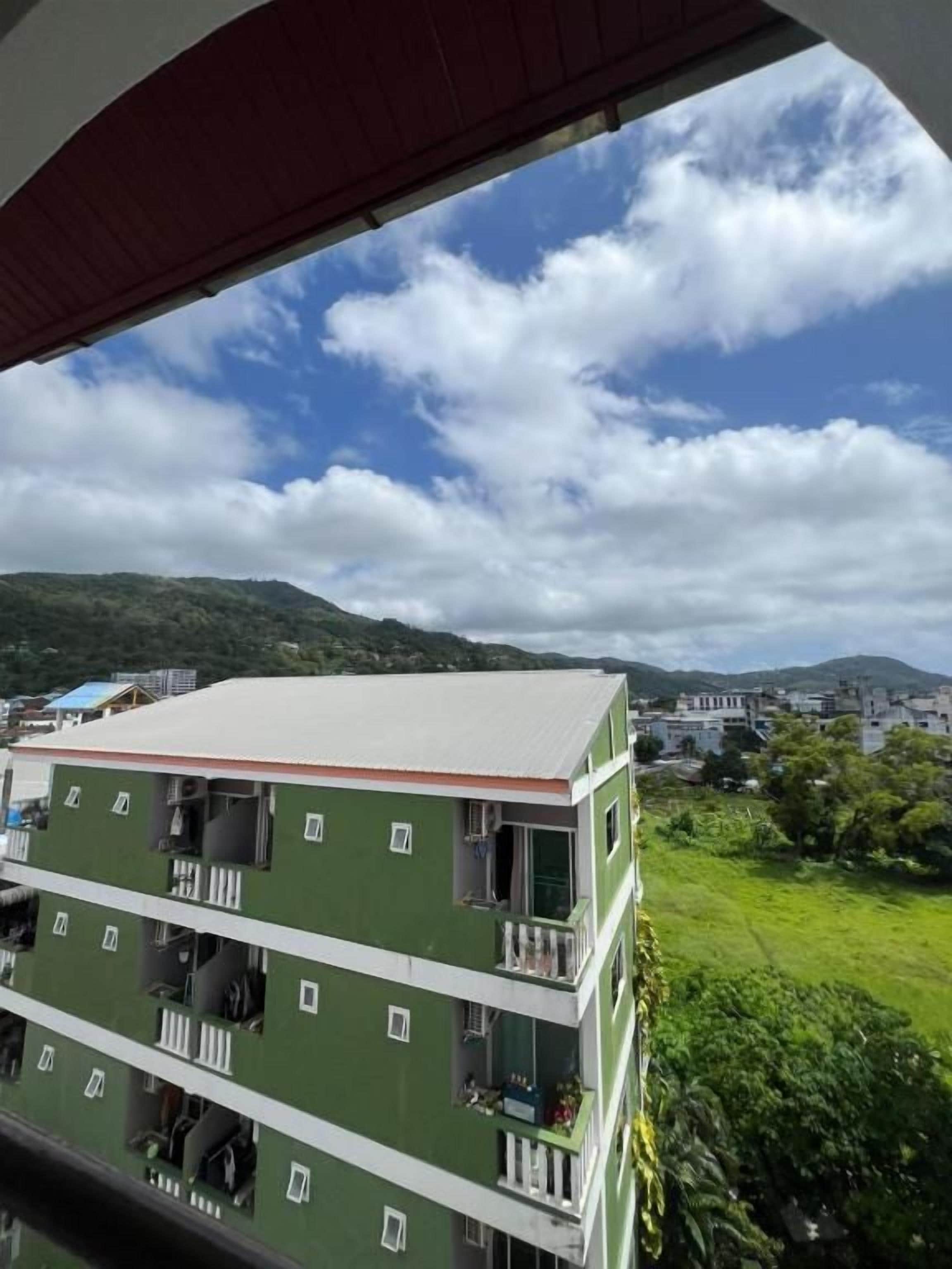 Un edificio de varios pisos con balcones, paredes exteriores verdes y vistas a un paisaje montañoso bajo un cielo parcialmente nublado.
