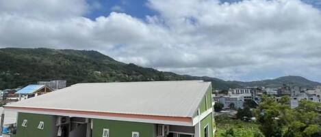 Un edificio de varios pisos con balcones, paredes exteriores verdes y vistas a un paisaje montañoso bajo un cielo parcialmente nublado.