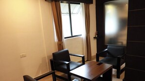 Un salon moderne avec un canapé, deux fauteuils et une table basse. On y trouve une fenêtre avec des rideaux, un téléviseur à écran plat fixé au mur et un sol carrelé.