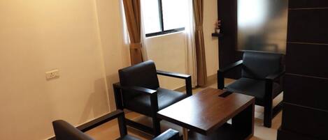 Una sala de estar moderna con un sofá, dos sillones y una mesa de centro. Hay una ventana con cortinas, un televisor de pantalla plana montado en la pared y un piso de baldosas.