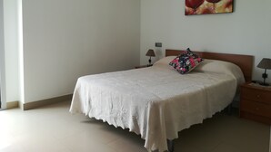 Un lit bien fait, recouvert d’une couvre-lit blanche et agrémenté d’un coussin à motifs floraux, une table de chevet avec une lampe et un tableau accroché au mur.