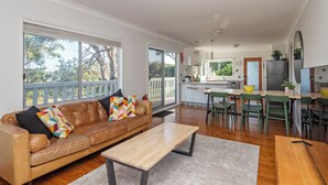 A modern living room with a leather sofa, a wooden coffee table, and a dining area with green chairs and a kitchen in the background.
