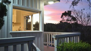 A wooden deck with a railing, a window with a view of the interior, and a sunset in the background.