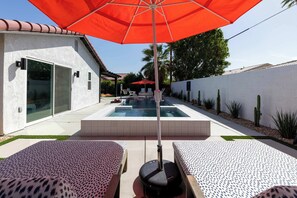 A pool area with two lounge chairs, a red umbrella, and a hot tub.