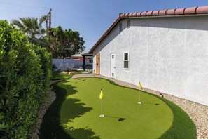 Ein gepflegter, künstlicher Rasenplatz für Golf mit einer Fahne, umgeben von einem weißen Gebäude mit roten Ziegeln.
