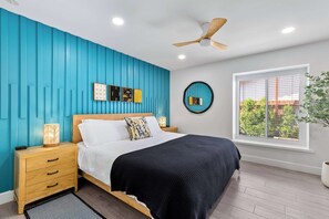 Un dormitorio con cama, mesita de noche, cuadro en la pared, ventilador de techo, ventana y planta en maceta.