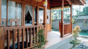 Una veranda de madera con un área de estar techada, una piscina pequeña y un jardín con plantas y una fuente.