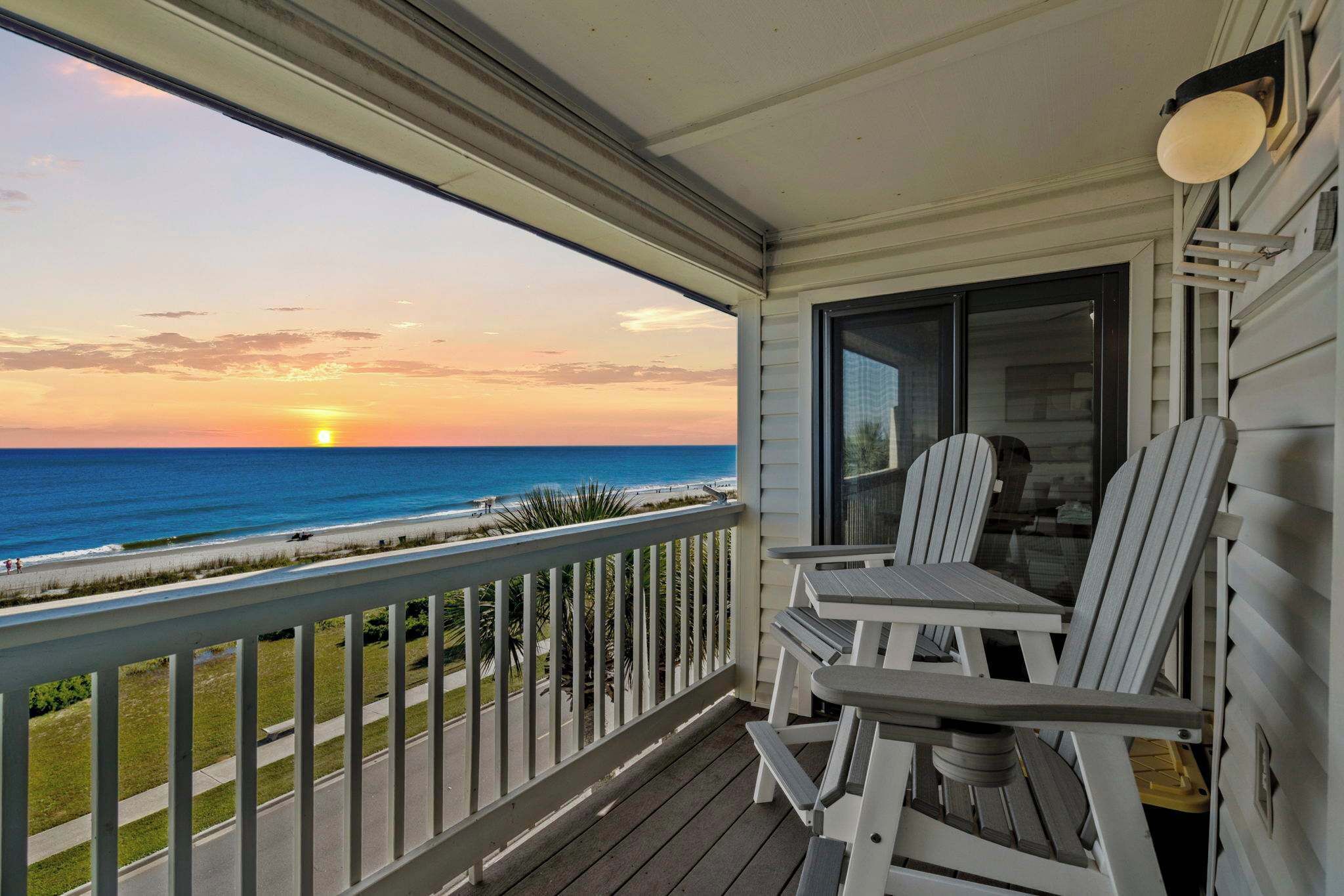 A balcony with a view of the ocean at sunset, featuring white Adirondack chairs and a table.