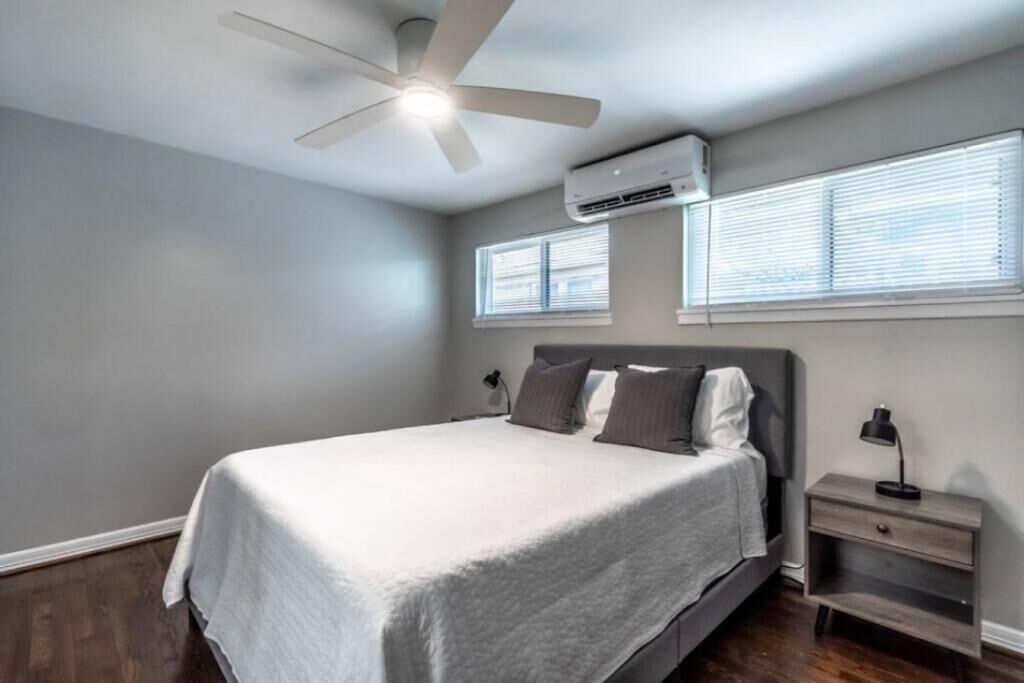 A bedroom with a bed, a ceiling fan, an air conditioner, a nightstand, and a window with blinds.