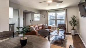 A modern living room with a brown leather sofa, a dining table with a potted plant, a ceiling fan, and a large window with curtains. - Stylish 1BR Urban Retreat (Houston)