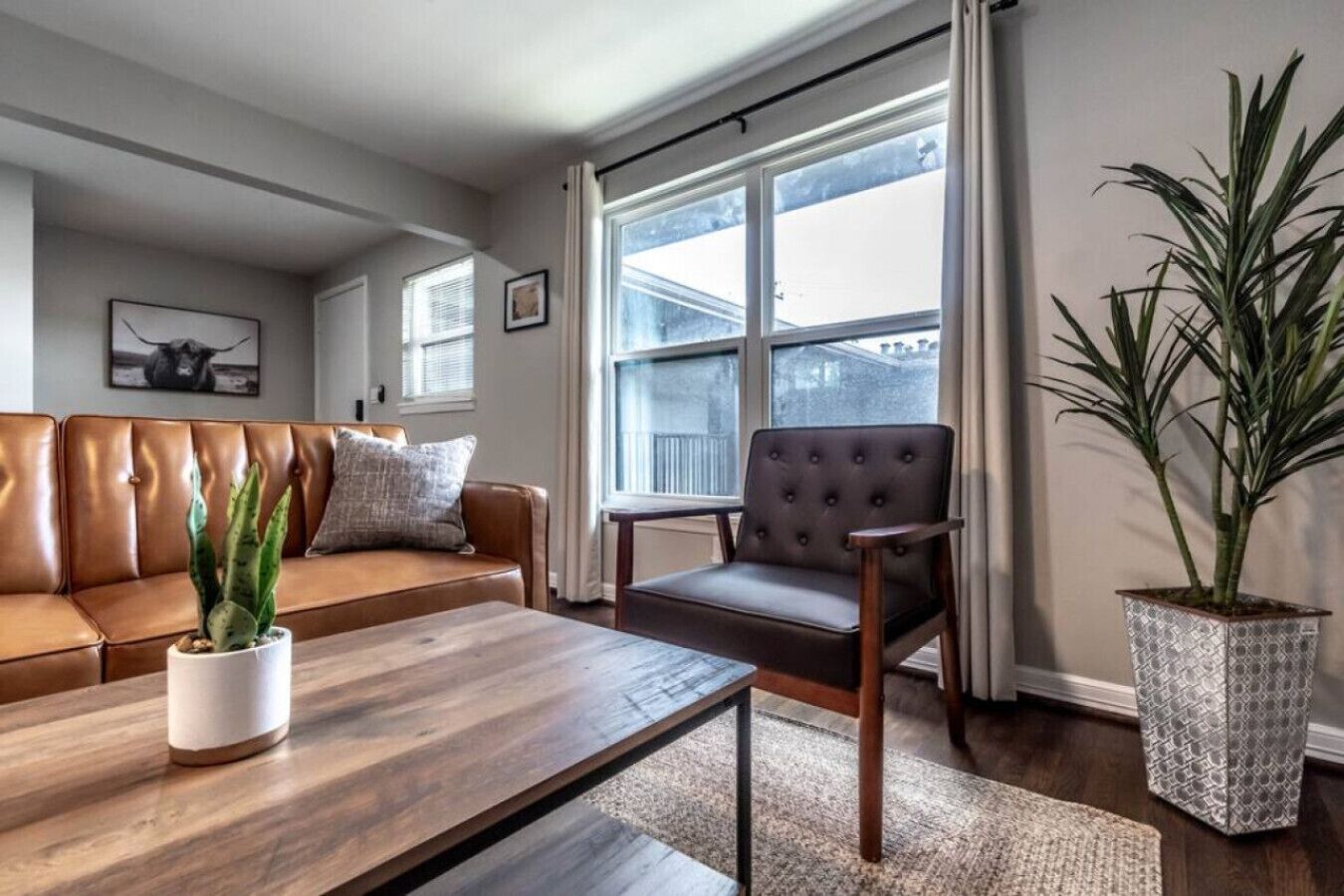 Een woonkamer met een bruine leren bank, een houten salontafel, een gestofte fauteuil en een groot raam met gordijnen.