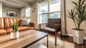 A living room with a brown leather sofa, a wooden coffee table, a grey armchair, and a large potted plant. - Stylish 1BR Urban Retreat (Houston)