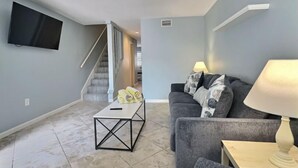 A living room with a grey sofa, a coffee table, a TV mounted on the wall, and a staircase leading to another room. - Stylish 1BR Urban Retreat (Houston)
