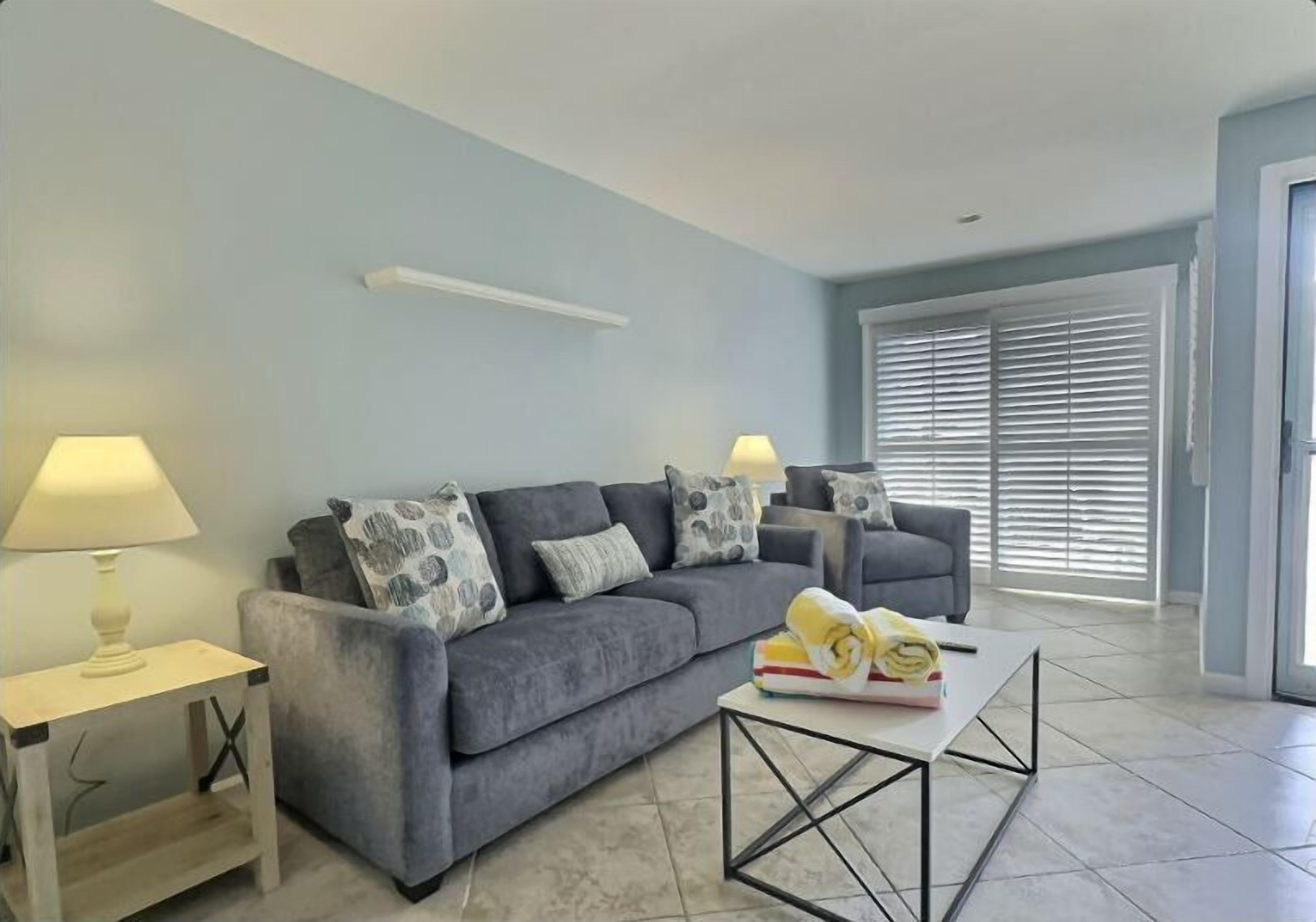 A living room with a grey sofa, two side tables with lamps, a coffee table, and a window with blinds.