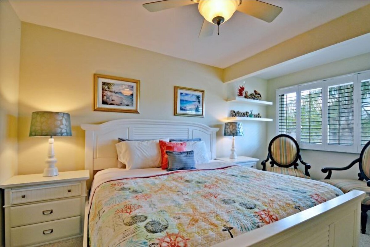 A bedroom with a bed, two chairs, a nightstand, a lamp, a ceiling fan, and a window with blinds.