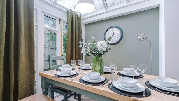 Ein Essbereich mit einem Holztisch, gedeckt für sechs Personen, einer grünen Vase mit Blumen, einer Wanduhr und einem Fenster mit Vorhängen.