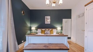 Ein Schlafzimmer mit dunkler Wand, einem Holz-Kopfteil, zwei Nachttischlampen, einem Bett mit Kissen, einem Nachttisch und einer hölzernen Bank.