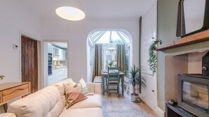 Ein gemütliches Wohnzimmer mit einer Couch, einem Esstisch und einem Fernseher. Ein Fenster mit Vorhängen, eine Pflanze und ein Kamin sind ebenfalls vorhanden.