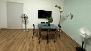 Un coin repas comprenant une table en verre, quatre chaises, une plante en pot, un ventilateur sur pied, un téléviseur fixé au mur et un ventilateur au sol.