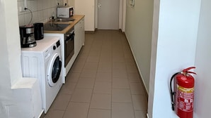 Un couloir étroit, avec une machine à laver, une machine à café et un four à micro-ondes sur le côté gauche. Une porte se trouve au bout du couloir.
