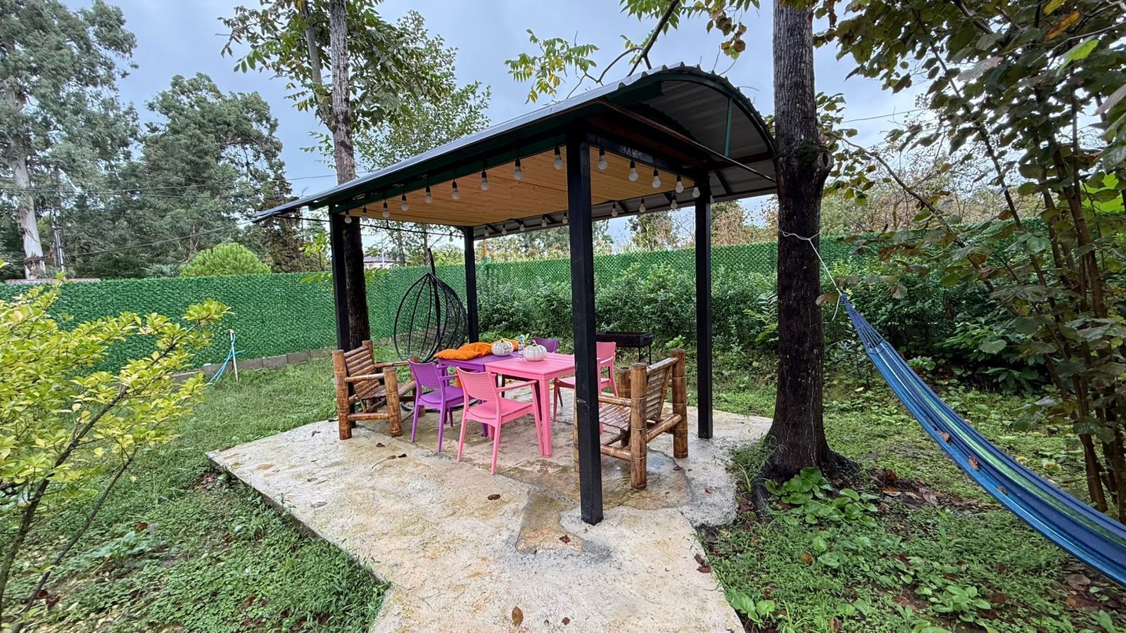 Un espace extérieur aménagé avec une table et des chaises, protégé par un auvent et entouré de verdure, avec un hamac à proximité.
