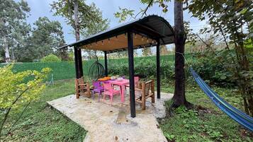 Een overdekt terras met een tafel en stoelen, omringd door groen en een hangmat.