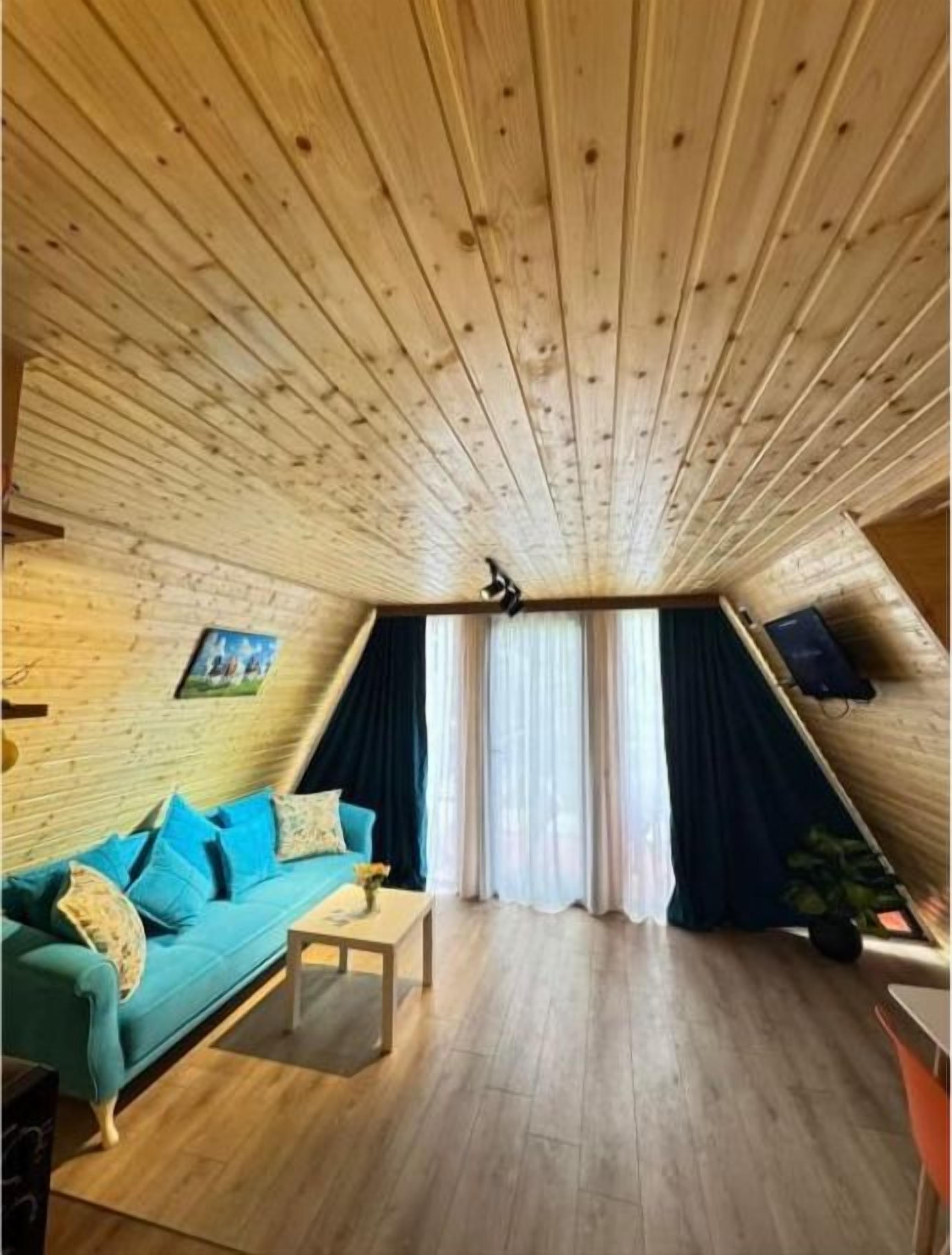 Acogedor cuarto ático con techo de madera, un sofá color esmeralda, una mesita y una ventana con cortinas.