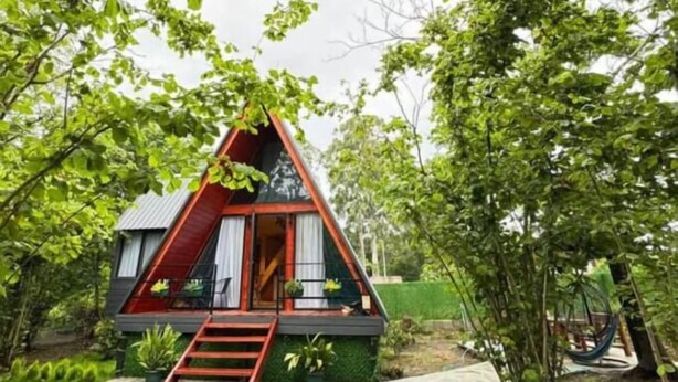 Une petite cabane triangulaire, avec un toit rouge, un escalier en bois menant à l’entrée, et entourée d’une végétation luxuriante.
