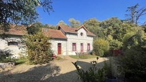Un bâtiment en pierre de deux étages, doté de volets rouges et d’un toit en pente, entouré d’une végétation luxuriante et d’arbres.