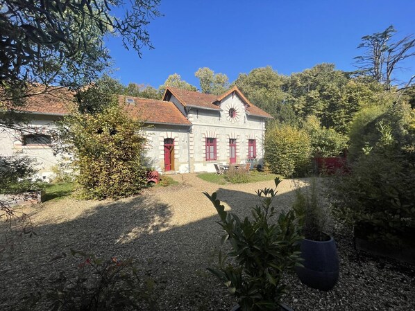 Un bâtiment en pierre de deux étages, doté de volets rouges et d’un toit en pente, entouré d’une végétation luxuriante et d’arbres.