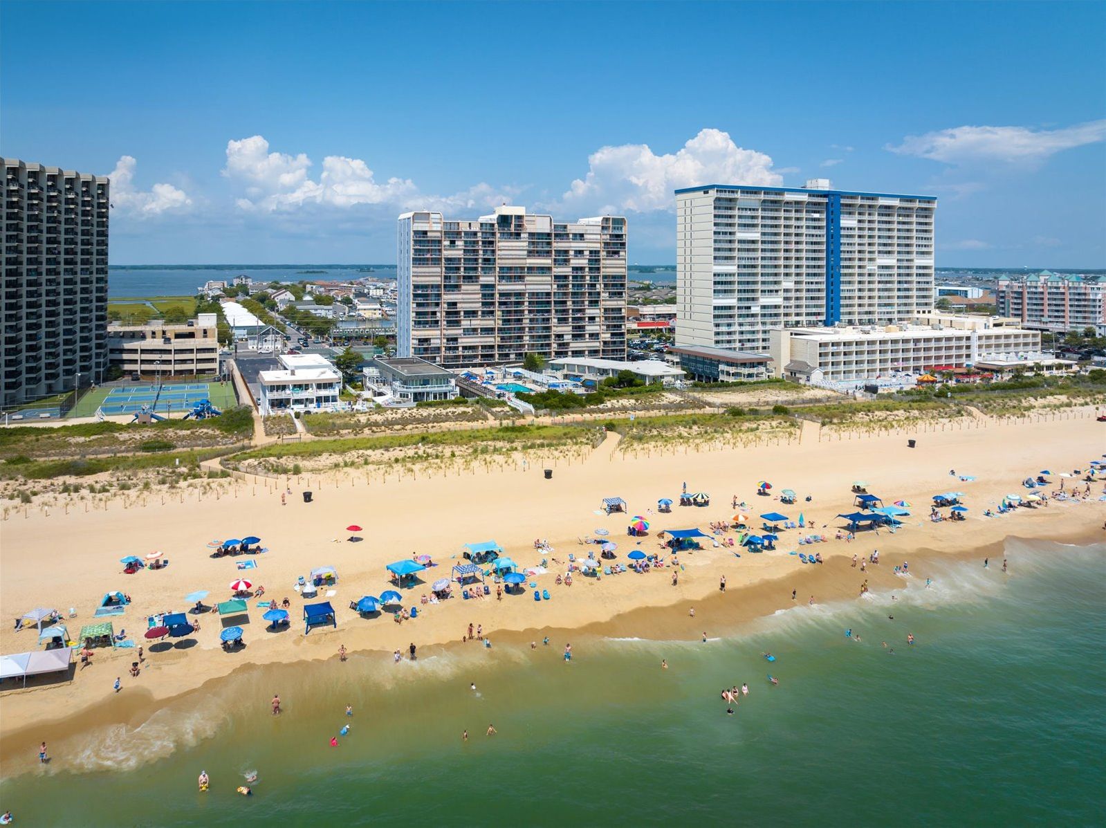Une plage bordée de nombreux parasols et fréquentée par de nombreuses personnes, entourée de grands hôtels et d’immeubles.