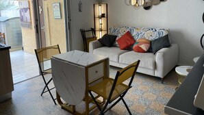 Een woonkamer met een bank, een salontafel en een eettafel met stoelen. Er hangt een spiegel aan de muur en een klein plankje met decoratieve objecten.