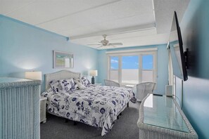 Quarto com cama, cadeira, mesa, ventoinha no teto, janela e televisão.