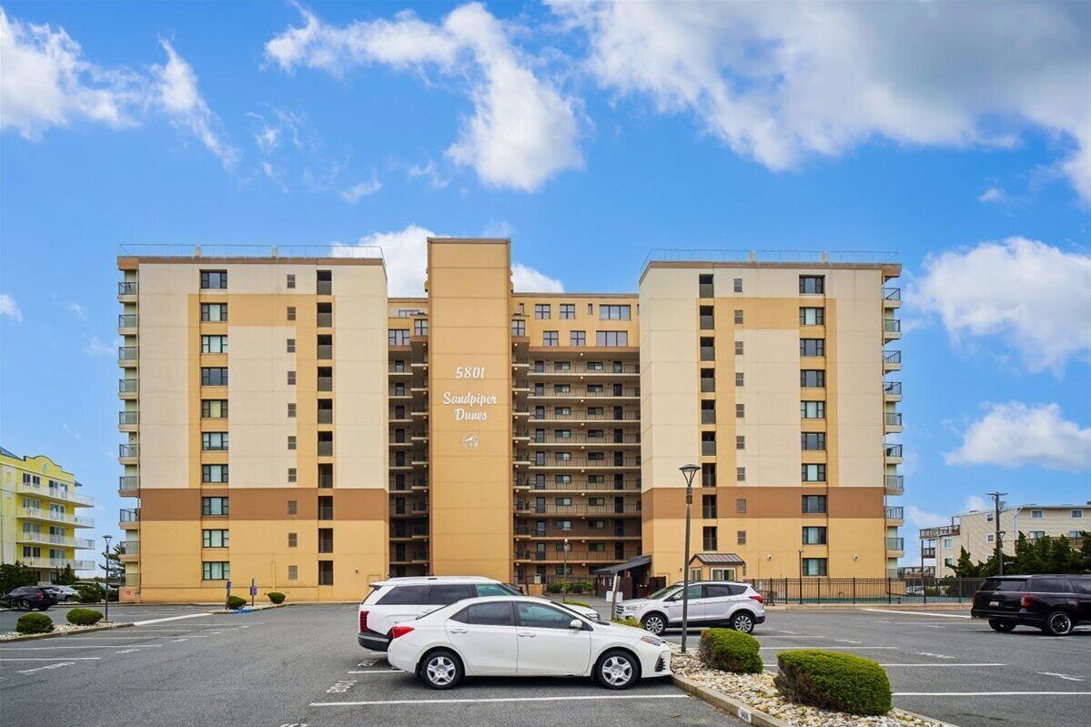 A multi-story hotel with a central entrance, parking area, and a clear sky.